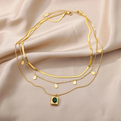 A gold-tone layered necklace displayed on a silky beige fabric. The necklace features three delicate chains: a flat snake chain, a chain with small gold teardrop charms, and a dainty chain with a square pendant containing a green gemstone-like detail. The necklace has an adjustable lobster clasp closure with an extender chain.