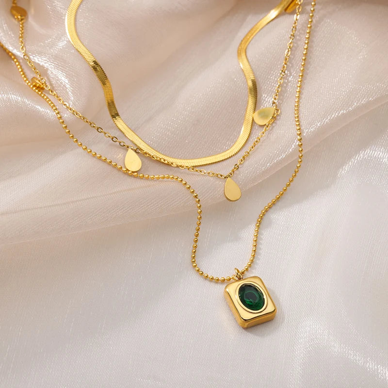 A gold-tone layered necklace displayed on a silky beige fabric. The necklace features three delicate chains: a flat snake chain, a chain with small gold teardrop charms, and a dainty chain with a square pendant containing a green gemstone-like detail. The necklace has an adjustable lobster clasp closure with an extender chain.